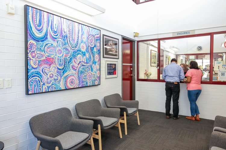 2 adults standing at the office counter being assisted by a member of the office staff. There are 3 chairs under a blue aboriginal painting in the foreground.