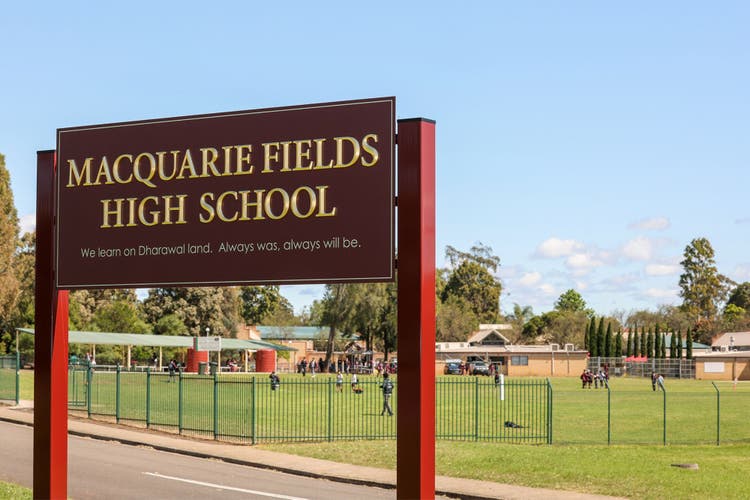 Maroon school sign with gold lettering with a view of the school oval behind and school buildings in the distance