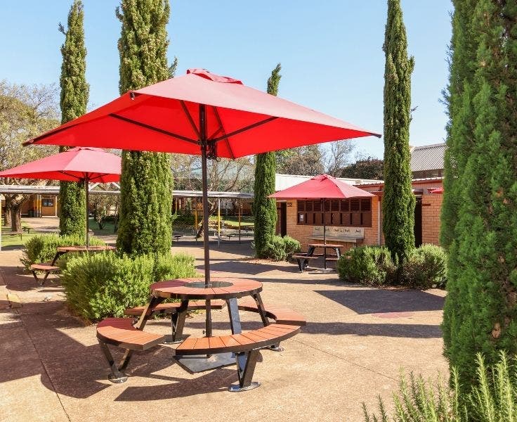 3 large square red umbrellas each above a round picnic table and seats. Located in a garden with tall thin pencil pines